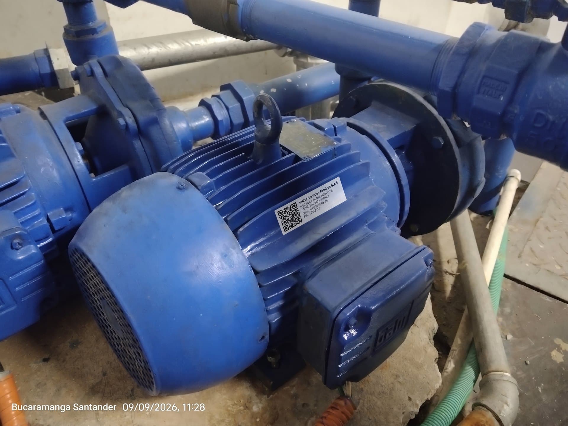 Motor eléctrico azul de una motobomba en un cuarto de bombas, con una etiqueta de código QR y los datos del equipo pegada sobre la carcasa.